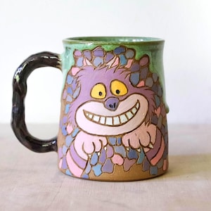 Puede incluir: Una taza de cerámica con una criatura morada pintada a mano que muestra una amplia sonrisa con dientes y ojos amarillos. La taza tiene un borde verde, una base marrón texturizada y un asa oscura y retorcida. La criatura está rodeada de formas azules.