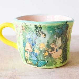 May include: A whimsical ceramic mug with a yellow handle and a light green exterior. The mug features a colorful illustration of fairies, butterflies, and mushrooms in a garden setting. The interior is a soft pink.