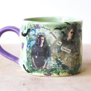 Puede incluir: Una taza de cerámica con exterior verde claro y asa morada. La taza presenta imágenes de un hombre, mariposas y murciélagos, con el texto "It's always gonna be Stefan."