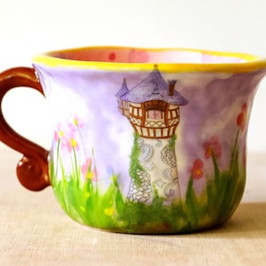 Puede incluir: Una taza de cerámica caprichosa con un asa marrón y un diseño colorido. La taza presenta una ilustración de una torre con un tejado morado, sobre un fondo lavanda con flores y hierba verde. El borde es amarillo.