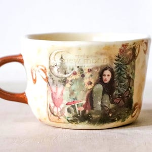 Könnte beinhalten: Ein cremefarbener Keramikbecher mit glänzendem braunem Henkel, bedruckt mit einer Grafik einer Person in einer Waldlandschaft, einer Mondsichel und dem Text "Twilight New Moon". Das Design zeigt zudem Schmetterlinge, Blumen und Pilze.