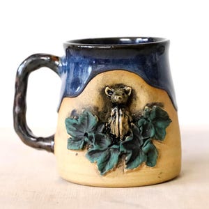 Puede incluir: Una taza de cerámica hecha a mano con borde y asa azul oscuro. La taza presenta un cuerpo beige con un diseño esculpido de un pequeño animal y hojas verdes. El asa es de color marrón oscuro y texturizada.