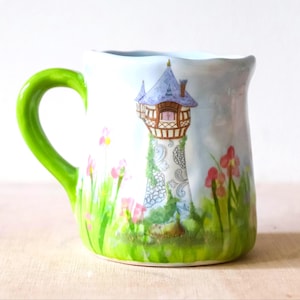 Puede incluir: Taza de cerámica con asa verde y un diseño de torre caprichoso. La torre tiene un tejado azul y detalles marrones, rodeada de flores rosas y hierba verde. La taza es blanca con un borde festoneado.