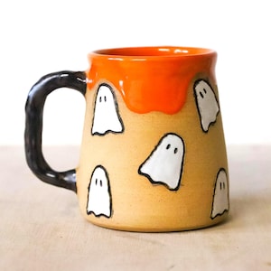 Puede incluir: Taza de cerámica con asa negra y cuerpo beige. El borde superior es naranja, con ilustraciones de fantasmas blancos delineadas en negro. La taza es un artículo temático de Halloween.