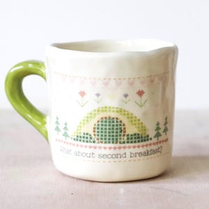 May include: A ceramic mug with a green handle and a cream-colored body. The mug features a cross-stitch design of a green and yellow arched structure, trees, and flowers. The text "What about second breakfast?" is printed on the front.
