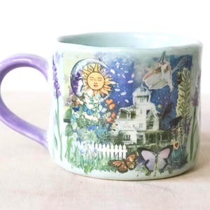 Könnte beinhalten: Ein hellblauer Keramikbecher mit violettem Henkel und einem Collage-Druck, der ein weißes Haus, Sonne, Mond, Schmetterlinge und florale Elemente zeigt. Das Design umfasst einen weißen Lattenzaun und verschiedene Gartenpflanzen um das Haus.