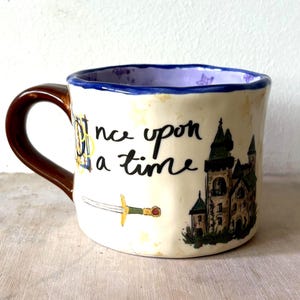 Puede incluir: Una taza de cerámica con exterior color crema que presenta el texto escrito a mano en negro 'once upon a time', una pequeña ilustración de una espada y un castillo detallado. La taza tiene un asa de color marrón oscuro y borde interior morado.
