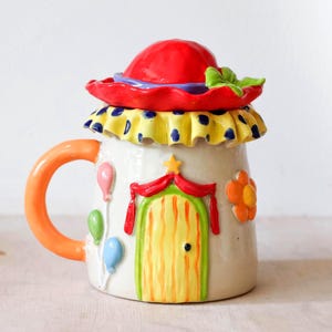 Könnte beinhalten: Ein Keramikbecher mit einem Deckel in Form eines roten Hutes. Der Becher hat einen gelben Rüschenrand, einen orangefarbenen Henkel und ein Reliefdesign mit einer gelben Tür, roten Vorhängen, einer orangefarbenen Blume und drei Luftballons.
