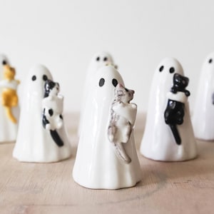 Puede incluir: Un conjunto de figuras de fantasmas de cerámica blanca, cada una sosteniendo un pequeño gato. Los gatos son de varios colores, incluyendo negro, gris y amarillo. Los fantasmas tienen ojos de puntos negros. Decoraciones de Halloween.