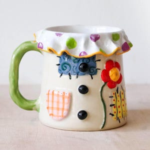 May include: A ceramic mug with a cream-coloured base, featuring a green handle and a ruffled rim decorated with purple and green polka dots. The mug is adorned with a red flower, a blue square patch, an orange checkered patch, and black buttons.