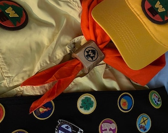 May include: A yellow scout hat with a brown patch featuring a tree and the letter W. A red neckerchief with a wooden patch featuring a tree and the letter W. A yellow shirt with a brown patch featuring a tree and the letter W. A black bag with various colorful patches featuring different designs.