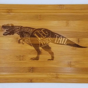 May include: A brown bamboo cutting board with a dinosaur silhouette engraved on it. The dinosaur is labeled with different cuts of meat, including "Neck", "Shoulder", "Breast", "Leg", "Thigh", and "Tail".
