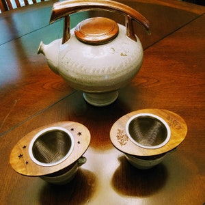 May include: A white ceramic teapot with a brown lid and wooden handle, along with two wooden tea infusers. One infuser has a star design and the other has a floral design.