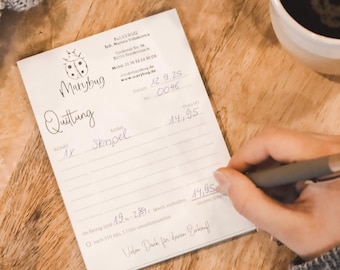 May include: A white paper receipt with the text Marybug and Quittung printed at the top, featuring a ladybug icon. The receipt includes handwritten details and a price of 14.95. A grey pen is positioned on the paper, which rests on a wooden surface.