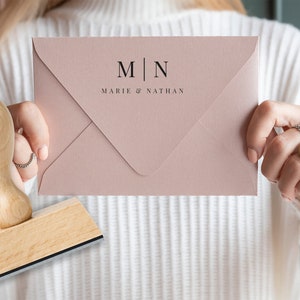 Könnte beinhalten: Ein blassrosa Umschlag mit den Initialen "M | N" und den Namen "Marie & Nathan" in Schwarz gedruckt. Ein Holzstempel und ein Ring mit Blattmuster sind ebenfalls zu sehen. Das Thema ist Hochzeit oder Schreibwaren.