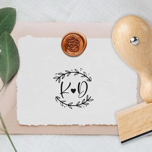Könnte beinhalten: Individueller Hochzeitsstempel mit Holzgriff, mit den Initialen "K & D" in einem Kranz-Design. Ein rosaförmiges Wachssiegel und Briefpapier sind ebenfalls zu sehen, ideal für personalisierte Einladungen.