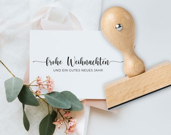May include: A wooden stamp with the words "frohe Weihnachten" and "und ein gutes neues Jahr" in black script. The stamp is next to a white card with the same text, decorated with pink flowers and eucalyptus leaves.