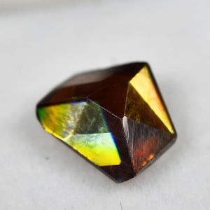 May include: A faceted gemstone with a deep brown base color and iridescent flashes of green and yellow light. The stone has a geometric, multi-sided cut and is displayed on a square white foam insert within a small black container.