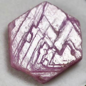 May include: A hexagonal pink gemstone with a flat, polished surface featuring distinct, intersecting linear patterns and geometric striations. The crystal displays a translucent, light pink hue with a reflective, slightly textured top surface.