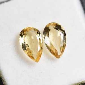 May include: Two pear-shaped, faceted gemstones with a warm, golden-yellow hue. The gems are displayed in a white box with a black border, creating a striking contrast. The stones are likely for jewelry making.