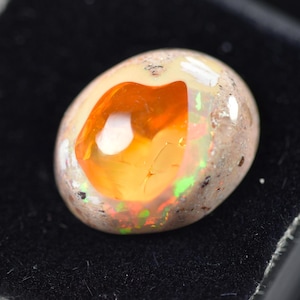 May include: Oval-shaped opal gemstone with a vibrant orange core and a speckled, light-colored exterior. The gemstone displays flashes of green and red, set against a black background.