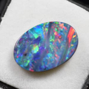 May include: An oval-shaped black opal gemstone featuring a dark base color with vibrant flashes of blue, green, and red light. The polished stone displays a multi-colored play-of-color effect across its smooth, rounded surface.