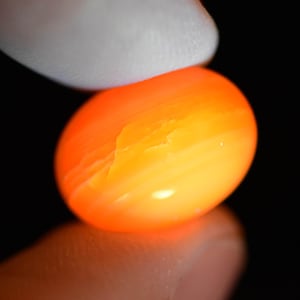 Op de afbeelding: Een ovale, oranje edelsteen met een gloeiend, doorschijnend uiterlijk. De edelsteen wordt vastgehouden tussen een wit gehandschoende vinger en een vingertop tegen een zwarte achtergrond. Het oppervlak van de steen heeft subtiele texturen en kleurvariaties.