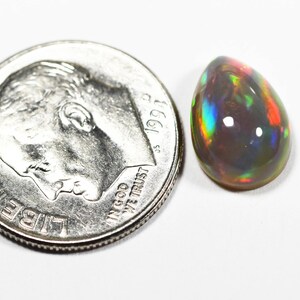 May include: A teardrop-shaped black opal gemstone displaying iridescent flashes of red, green, and blue, positioned next to a United States dime for scale to show the small size of the stone relative to the 0.705 inch diameter coin.