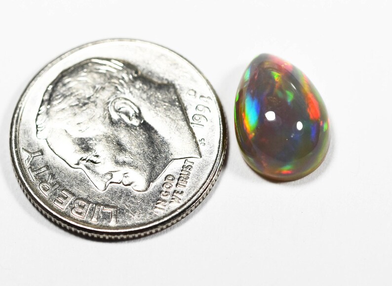 May include: A teardrop-shaped black opal gemstone displaying iridescent flashes of red, green, and blue, positioned next to a United States dime for scale to show the small size of the stone relative to the 0.705 inch diameter coin.