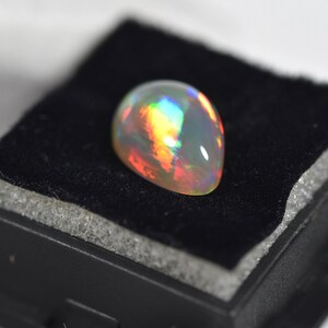 May include: A smooth, pear-shaped opal gemstone displaying a vibrant play-of-color with flashes of red, orange, yellow, green, and blue against a translucent, milky white base. The polished stone features a domed cabochon cut surface.
