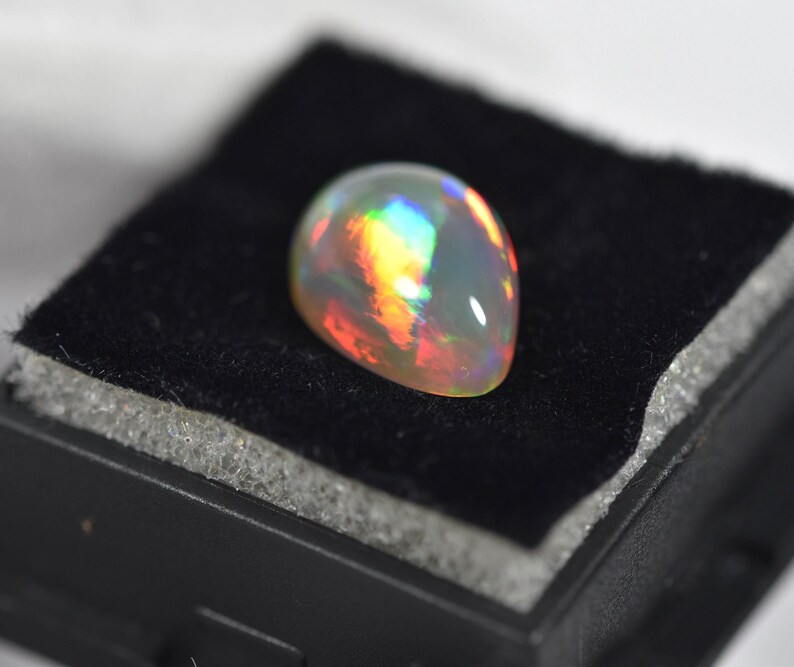 May include: A smooth, pear-shaped opal gemstone displaying a vibrant play-of-color with flashes of red, orange, yellow, green, and blue against a translucent, milky white base. The polished stone features a domed cabochon cut surface.