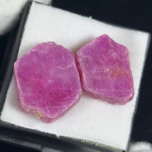 May include: Two raw, hexagonal-shaped ruby crystals with a vibrant, saturated pinkish-red hue. The stones feature a natural, unpolished, and textured surface with visible crystalline structures, displayed side-by-side in a small, square box.