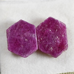 May include: Two raw, hexagonal-shaped rubies with a deep magenta-pink color and a textured, opaque surface. The stones exhibit natural crystalline structures with visible inclusions and facets across their flat, top-facing surfaces.