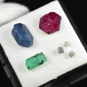 以下が含まれることがあります： 大きなブルーサファイア、深い赤色のルビー、長方形の緑色のエメラルド、そして3つの小さな半透明の未加工ダイヤモンドを含む原石のコレクション。黒い収納ケース内の白いフォームディスプレイパッドの上に配置されています。