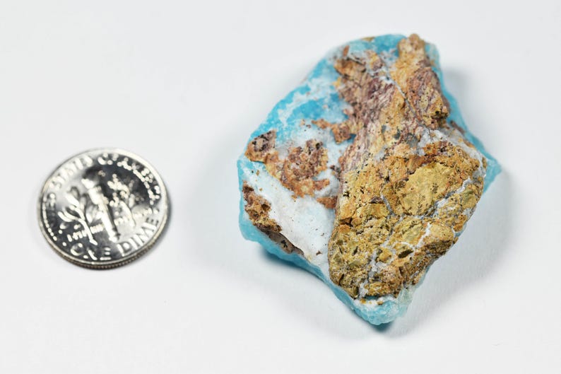 May include: A piece of raw turquoise gemstone with a dime for size comparison. The turquoise has a light blue hue with areas of brown and tan matrix. The dime is silver and shows the profile of Franklin D. Roosevelt.