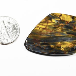May include: A polished, irregularly shaped gemstone with a mix of gold, blue, and brown hues. The stone is next to a US dime coin for size comparison. The gemstone's surface has a glossy finish and a unique pattern.
