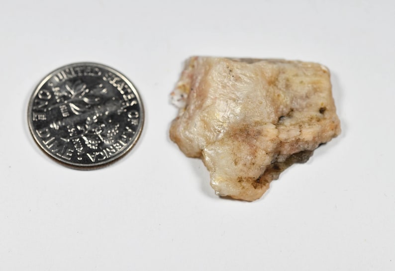 May include: A close-up of a raw, beige-colored mineral specimen next to a U.S. dime coin. The mineral has a rough, uneven texture with hints of pink and brown. The coin is silver and shows the profile of a person.