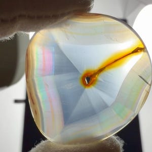 May include: A translucent, circular agate slice featuring a central amber-colored inclusion with a radiating pattern. The stone displays soft white and blue tones with iridescent, rainbow-like bands along the curved edges, backlit to reveal internal structure.