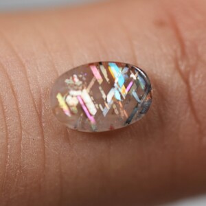 May include: An oval-shaped gemstone with a clear base and internal iridescent patterns of pink, blue, and yellow. The gemstone has a polished surface and a geometric design, reflecting light.