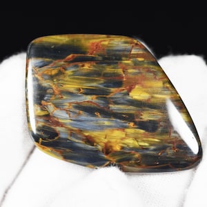 May include: A polished gemstone with a unique pattern of swirling colors. The stone features shades of gold, blue, and red, creating a striking visual effect. The gemstone is held against a white background.