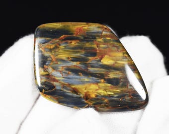 May include: A polished gemstone with a unique pattern of swirling colors. The stone features shades of gold, blue, and red, creating a striking visual effect. The gemstone is held against a white background.