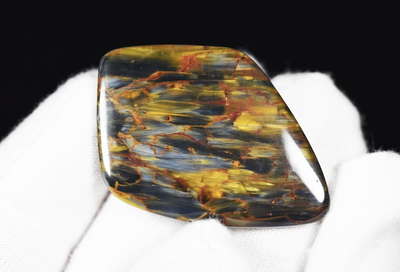 May include: A polished gemstone with a unique pattern of swirling colors. The stone features shades of gold, blue, and red, creating a striking visual effect. The gemstone is held against a white background.