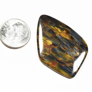 May include: A polished pietersite gemstone with a mix of blue, gold, and brown colors. The stone is irregularly shaped and lies next to a U.S. dime for size comparison.