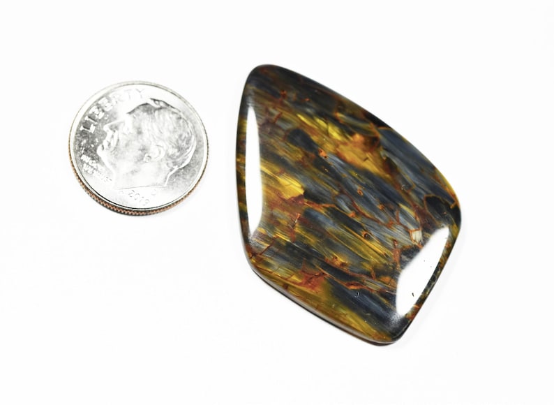 May include: A polished pietersite gemstone with a mix of blue, gold, and brown colors. The stone is irregularly shaped and lies next to a U.S. dime for size comparison.