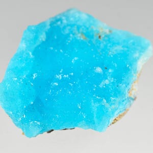 May include: A vibrant, rough-cut turquoise gemstone with a crystalline structure. The stone displays a bright, saturated blue color with a slightly uneven surface. The edges have a lighter, more opaque appearance, revealing a natural, unpolished texture.