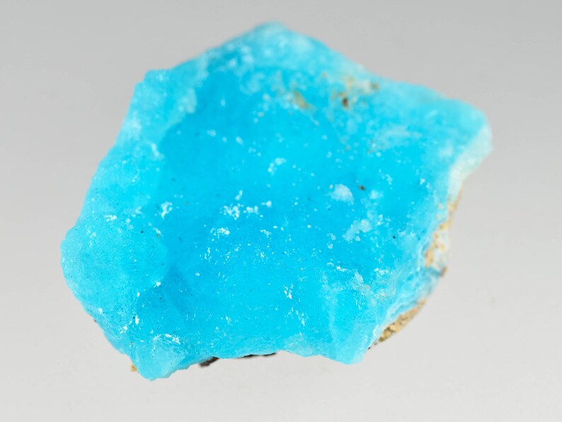 May include: A vibrant, rough-cut turquoise gemstone with a crystalline structure. The stone displays a bright, saturated blue color with a slightly uneven surface. The edges have a lighter, more opaque appearance, revealing a natural, unpolished texture.