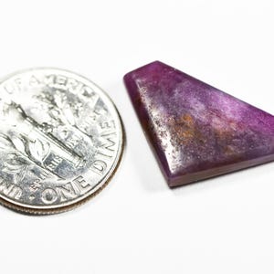 May include: A polished, irregular trapezoidal-shaped purple gemstone with visible dark inclusions and a smooth surface, positioned next to a United States one dime coin for scale to show its relative size.