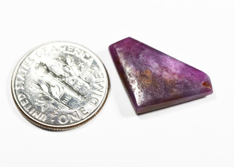 May include: A polished, irregular trapezoidal-shaped purple gemstone with visible dark inclusions and a smooth surface, positioned next to a United States one dime coin for scale to show its relative size.