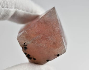 May include: A polished, multifaceted pink quartz crystal point with visible internal inclusions and small black mineral deposits at the base. The stone features a translucent, pale rose hue with a geometric, pointed top and a rougher, textured bottom.