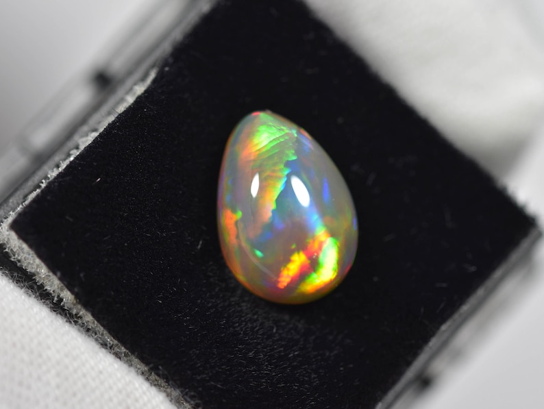 May include: A pear-shaped opal gemstone featuring a translucent white base with vibrant flashes of red, orange, yellow, green, and blue play-of-color. The smooth, polished cabochon displays a high-domed surface that reflects light across its surface.